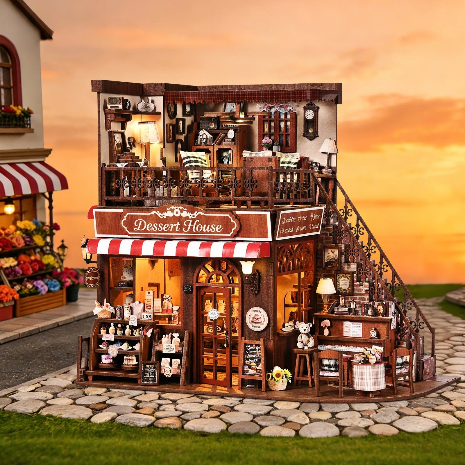 Maquette de pâtisserie miniature "Dessert House" illuminée, exposée devant un décor extérieur de coucher de soleil sur une rue pavée avec une boutique de fleurs en arrière-plan.