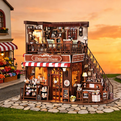 Maquette de pâtisserie miniature "Dessert House" illuminée, exposée devant un décor extérieur de coucher de soleil sur une rue pavée avec une boutique de fleurs en arrière-plan.