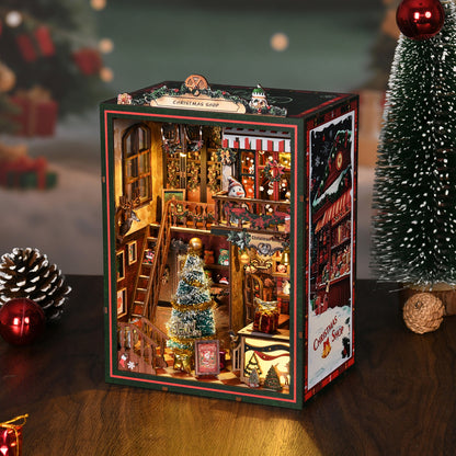 Serre-livres miniature DIY Book Nook 'Christmas Shop' en bois, décoré pour les fêtes. La scène intérieure comprend un sapin de Noël scintillant avec une étoile dorée, un bonhomme de neige sur un balcon, et une boutique remplie de cadeaux miniatures. L'ensemble est baigné dans une lumière LED jaune chaleureuse, créant une ambiance de village de Noël magique avec des détails rouges et verts festifs.