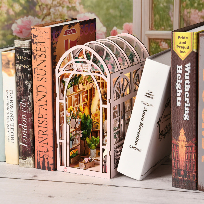 Book Nook 'Mini Greenhouse' installé sur une étagère entre des livres, montrant une serre miniature rose avec éclairage LED chaleureux, étagères de plantes et détails floraux.