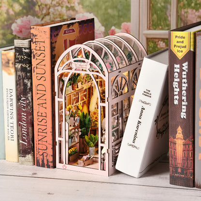 Book Nook 'Mini Greenhouse' installé sur une étagère entre des livres, montrant une serre miniature rose avec éclairage LED chaleureux, étagères de plantes et détails floraux.