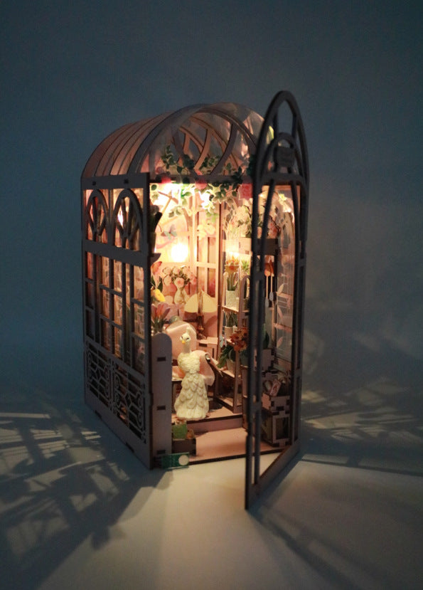 Book Nook 'Mini Greenhouse' en bois rose illuminé dans l'obscurité, montrant une serre miniature avec une porte ouverte, un paon blanc décoratif et des détails de fleurs et plantes.