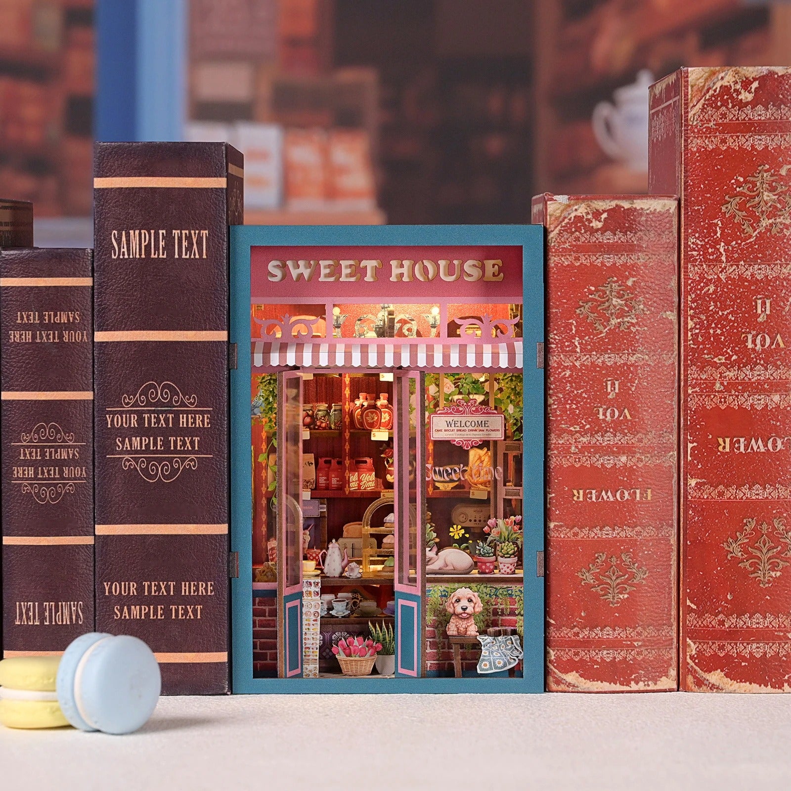 Kit DIY Book Nook Sweet House inséré entre des livres anciens sur une étagère. Maquette de pâtisserie miniature en bois avec éclairage chaleureux, fleurs, et vitrine détaillée. Décoration de bibliothèque cosy et romantique.