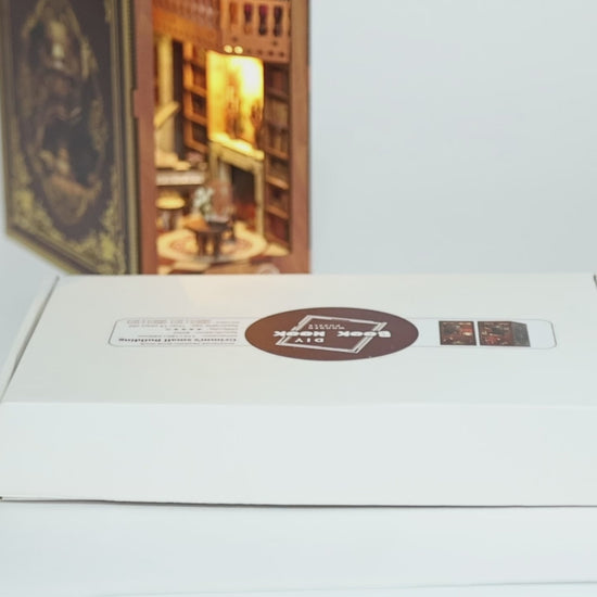 Présentation du kit DIY Book Nook 'Grimm’s Small Building'. La vidéo montre le déballage de la boîte, les pièces en bois découpées au laser, le manuel d'assemblage et le résultat final : une bibliothèque miniature rétro éclairée avec un piano, un escalier et des étagères de livres, parfaitement intégrée entre de vrais livres sur une étagère.