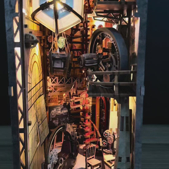 Vidéo de présentation d'une maquette Book Nook animée au style steampunk intitulée "Uncle Brian's Warehouse". La caméra explore l'intérieur détaillé en bois, révélant une montgolfière qui oscille doucement, des engrenages en mouvement et une grande roue mécanique. L'éclairage LED chaleureux met en relief les escaliers en colimaçon, un projecteur de cinéma miniature et une moto rétro. Cette décoration de bibliothèque crée une ambiance industrielle vintage fascinante.