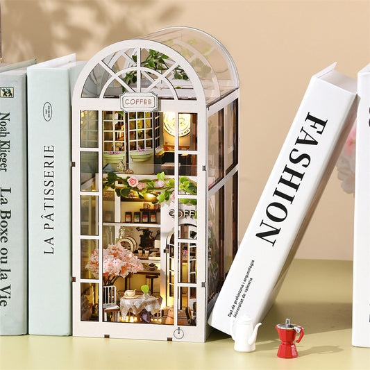 Serre-livres décoratif miniature (book nook) en bois blanc représentant un café à deux étages sous une verrière arrondie. Le diorama est niché entre des livres sur une étagère et est illuminé par une lumière chaleureuse. On aperçoit à l'intérieur des tables, des plantes grimpantes, des fleurs roses et un comptoir. À l'extérieur, une petite cafetière rouge et un pot à lait blanc sont posés devant la structure.