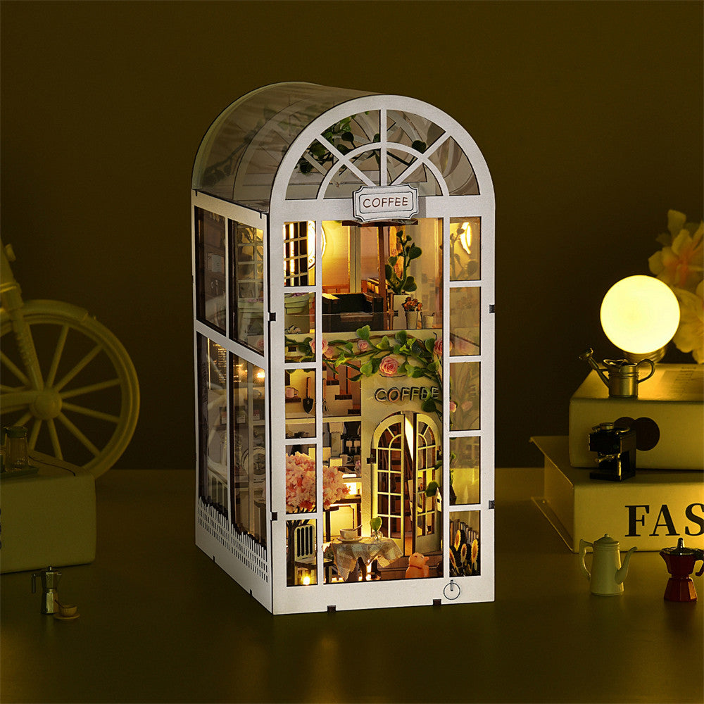 Un serre-livres miniature (book nook) en bois blanc représentant un café à deux étages au design élégant. La structure possède une grande verrière arrondie et est entièrement illuminée de l'intérieur par une lumière LED jaune. On distingue de nombreux détails comme des tables de café, des plantes fleuries roses et un comptoir. La maquette est mise en scène dans une ambiance tamisée, posée à côté d'une lampe ronde et de petits accessoires de cuisine miniatures.